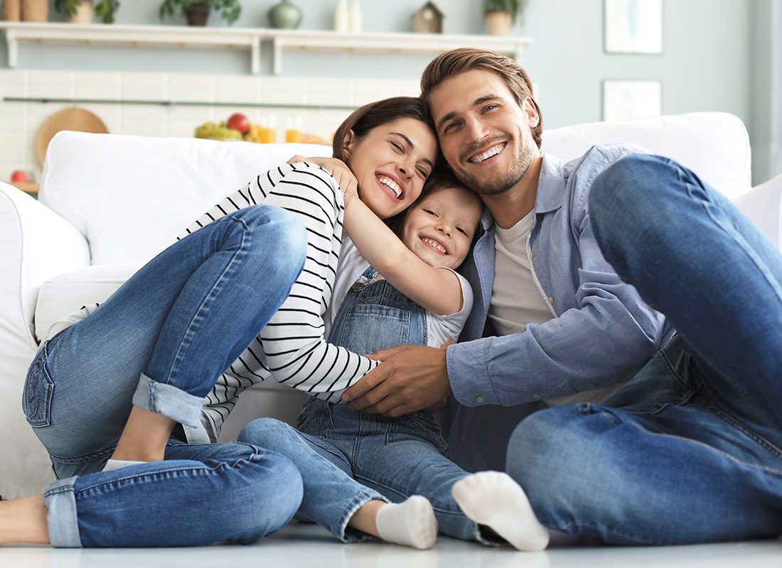 Personal Insurance - Closeup Portrait of Young Parents Hugging Their Daughter as They Have Fun Spending Time Together at Home Sitting on the Sofa